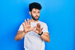 Young arab man with beard wearing casual white t shirt moving away hands palms showing refusal and denial with afraid and disgusting expression. stop and forbidden. 