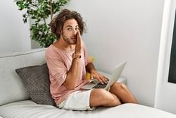 Young hispanic man sitting on the sofa at home using laptop hand on mouth telling secret rumor, whispering malicious talk conversation 