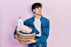 Handsome hipster young man holding laundry basket and detergent bottle smiling looking to the side and staring away thinking. 