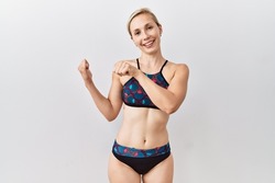 Young blonde woman wearing sporty bikini over isolated background pointing to the back behind with hand and thumbs up, smiling confident 