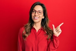 Young latin woman wearing casual clothes and glasses with a big smile on face, pointing with hand and finger to the side looking at the camera. 