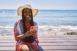 Young african american tourist woman using smartphone sitting on the bench at the beach.