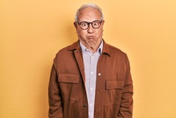 Senior man with grey hair wearing casual jacket and glasses puffing cheeks with funny face. mouth inflated with air, crazy expression. 