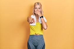 Beautiful blonde sports woman wearing workout outfit laughing at you, pointing finger to the camera with hand over mouth, shame expression 