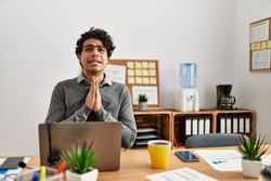 Young hispanic man wearing business style sitting on desk at office begging and praying with hands together with hope expression on face very emotional and worried. begging. 