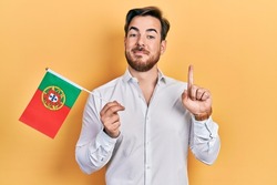 Handsome caucasian man with beard holding portugal flag smiling with an idea or question pointing finger with happy face, number one 