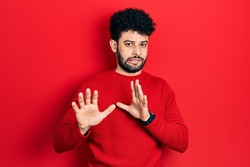 Young arab man with beard wearing casual red sweater disgusted expression, displeased and fearful doing disgust face because aversion reaction. with hands raised 