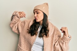 Young hispanic woman wearing wool sweater and winter hat showing arms muscles smiling proud. fitness concept. 