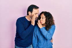 Middle age couple of hispanic woman and man hugging and standing together hand on mouth telling secret rumor, whispering malicious talk conversation 