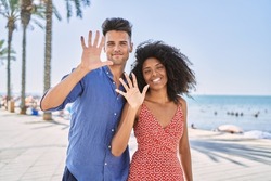 Young interracial couple outdoors on a sunny day showing and pointing up with fingers number ten while smiling confident and happy. 
