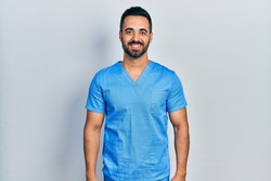 Handsome hispanic man with beard wearing blue male nurse uniform with a happy and cool smile on face. lucky person. 