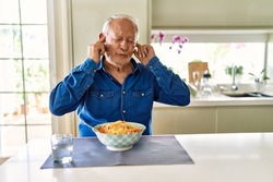 Senior man with grey hair eating pasta spaghetti at home covering ears with fingers with annoyed expression for the noise of loud music. deaf concept. 