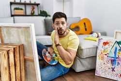 Young man with beard painting canvas at home hand on mouth telling secret rumor, whispering malicious talk conversation 