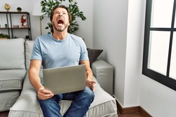 Handsome middle age man using computer laptop on the sofa angry and mad screaming frustrated and furious, shouting with anger. rage and aggressive concept. 
