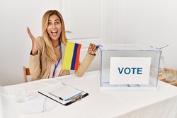 Blonde beautiful young woman at political campaign election holding colombia flag celebrating victory with happy smile and winner expression with raised hands 