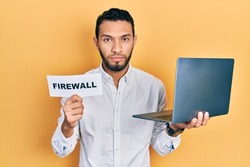 Hispanic man with beard holding computer laptop and firewall banner relaxed with serious expression on face. simple and natural looking at the camera. 