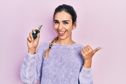 Young hispanic girl holding key of new car pointing thumb up to the side smiling happy with open mouth 