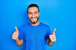 Hispanic man with beard wearing casual blue t shirt success sign doing positive gesture with hand, thumbs up smiling and happy. cheerful expression and winner gesture. 