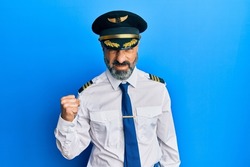 Middle age man with beard and grey hair wearing airplane pilot uniform angry and mad raising fist frustrated and furious while shouting with anger. rage and aggressive concept. 