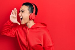 Beautiful hispanic woman with short hair listening to music using headphones shouting and screaming loud to side with hand on mouth. communication concept. 