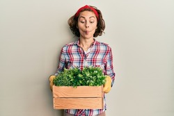 Young brunette woman wearing gardener clothes holding wooden plant pot making fish face with mouth and squinting eyes, crazy and comical. 