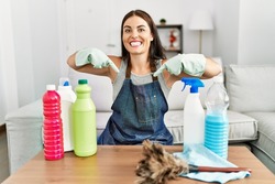 Young brunette woman wearing cleaner apron and gloves cleaning at home looking confident with smile on face, pointing oneself with fingers proud and happy. 