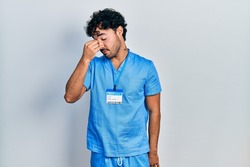 Young hispanic man wearing blue male nurse uniform tired rubbing nose and eyes feeling fatigue and headache. stress and frustration concept. 