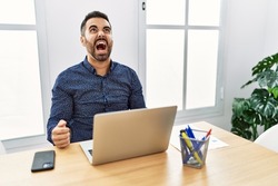 Young hispanic man with beard working at the office with laptop angry and mad screaming frustrated and furious, shouting with anger. rage and aggressive concept. 
