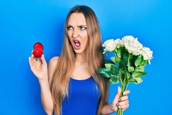 Young blonde girl holding bouquet of flowers and engagement ring angry and mad screaming frustrated and furious, shouting with anger. rage and aggressive concept. 