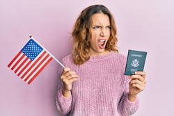 Young caucasian woman holding united states flag and passport angry and mad screaming frustrated and furious, shouting with anger. rage and aggressive concept. 