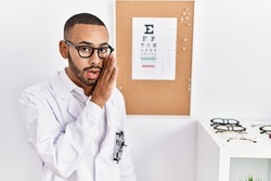 African american optician man standing by eyesight test hand on mouth telling secret rumor, whispering malicious talk conversation 