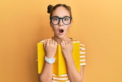 Beautiful brunette little girl wearing glasses and holding book afraid and shocked with surprise and amazed expression, fear and excited face. 
