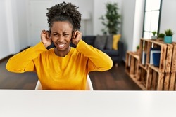 Young african american woman wearing casual clothes sitting on the table at home covering ears with fingers with annoyed expression for the noise of loud music. deaf concept. 