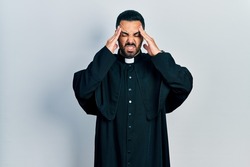 Handsome hispanic man with beard wearing catholic priest robe with hand on head, headache because stress. suffering migraine. 
