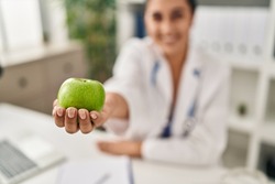 Young hispanic woman wearing dietitian uniform holding apple at clinic