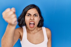 Young hispanic woman standing over blue background angry and mad raising fist frustrated and furious while shouting with anger. rage and aggressive concept. 