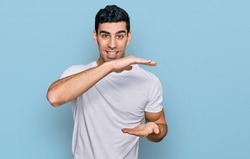 Handsome hispanic man wearing casual white t shirt gesturing with hands showing big and large size sign, measure symbol. smiling looking at the camera. measuring concept. 