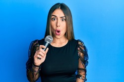Young hispanic woman singing song using microphone scared and amazed with open mouth for surprise, disbelief face 