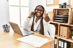 Black woman with braids working at the office speaking on the phone approving doing positive gesture with hand, thumbs up smiling and happy for success. winner gesture. 