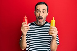 Middle age hispanic man holding ketchup and mustard bottle afraid and shocked with surprise and amazed expression, fear and excited face. 