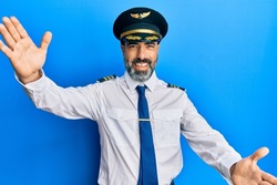 Middle age man with beard and grey hair wearing airplane pilot uniform looking at the camera smiling with open arms for hug. cheerful expression embracing happiness. 