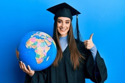 Beautiful brunette young woman wearing graduation robe holding world ball smiling happy and positive, thumb up doing excellent and approval sign 