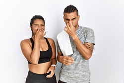 Young latin couple wearing sportswear standing over isolated background smelling something stinky and disgusting, intolerable smell, holding breath with fingers on nose. bad smell 
