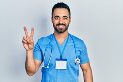Handsome hispanic man with beard wearing doctor uniform smiling with happy face winking at the camera doing victory sign. number two. 