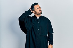 Handsome hispanic man with beard wearing catholic priest robe confuse and wondering about question. uncertain with doubt, thinking with hand on head. pensive concept. 