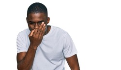 Young african american man wearing casual white t shirt smelling something stinky and disgusting, intolerable smell, holding breath with fingers on nose. bad smell 