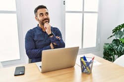 Young hispanic man with beard working at the office with laptop with hand on chin thinking about question, pensive expression. smiling and thoughtful face. doubt concept. 