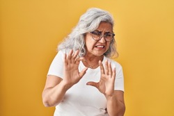 Middle age woman with grey hair standing over yellow background disgusted expression, displeased and fearful doing disgust face because aversion reaction. with hands raised 