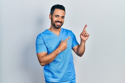 Handsome hispanic man with beard wearing blue male nurse uniform smiling and looking at the camera pointing with two hands and fingers to the side. 