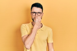 Young hispanic man wearing casual clothes and glasses smelling something stinky and disgusting, intolerable smell, holding breath with fingers on nose. bad smell 